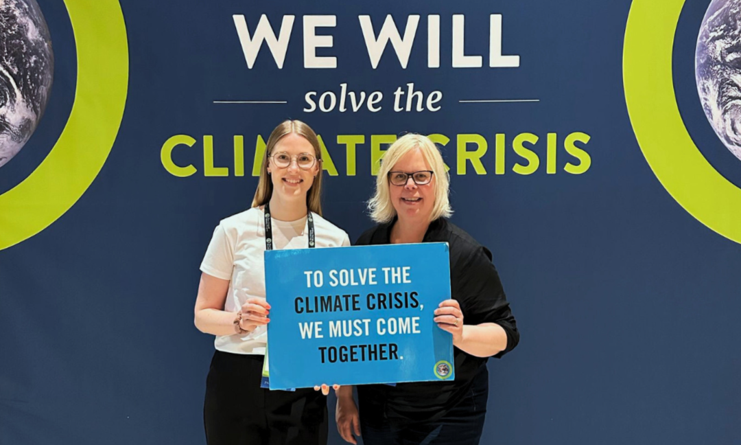 Laura and Hanna standing in front of The Climate Project's blue banderoll holding a light blue sign that reads: "To solve the climate crisis, we must come together."