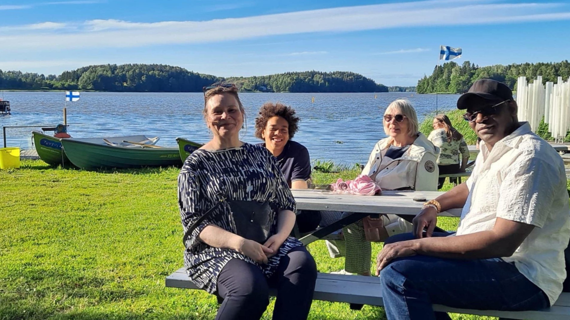 Nelihenkinen perhe hymyilee kameralle järven rannalla.